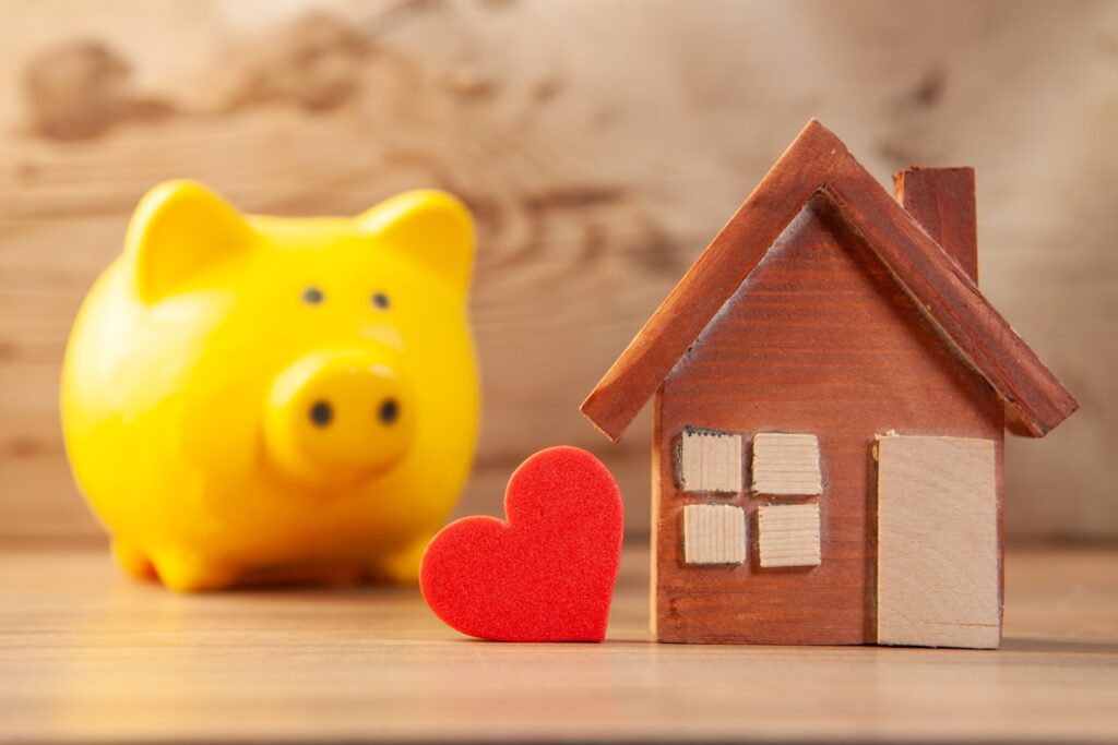 House, piggy bank and red heart on wooden background. Mortgage concept